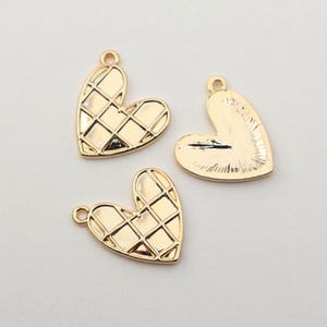 May include: Three gold-colored heart-shaped charms. Two charms have a grid pattern on the front, while the third charm shows the back with a smooth surface. Each charm has a small loop at the top for attaching to jewelry.