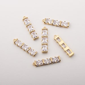 Puede incluir: Componentes de joyería rectangulares dorados con piedras rectangulares transparentes. Cada componente tiene un lazo en cada extremo para la fijación. Los componentes están dispuestos sobre una superficie blanca.