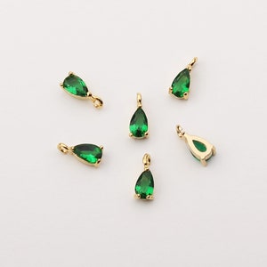 May include: Several gold-colored pendants with a teardrop-shaped, emerald-green gemstone. Each pendant has a small loop at the top for attaching to a chain or other jewelry component. The gemstones are faceted and catch the light.