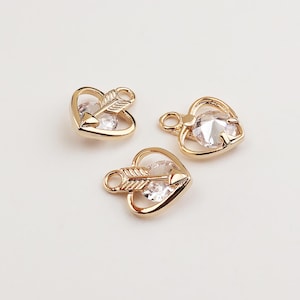 May include: Three gold-toned metal charms shaped like hearts, each featuring a central clear faceted gemstone and a decorative arrow motif across the centre. Each charm includes a small circular loop at the top for attachment to jewellery.