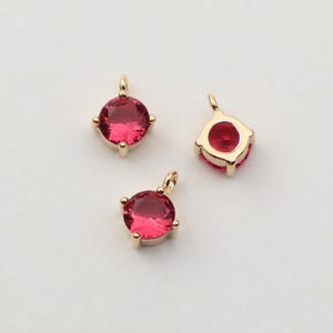 May include: Three gold-colored pendants with a round, deep pink gemstone. Each pendant has a small loop at the top for hanging. The gemstones are set in a gold-colored metal with four prongs.