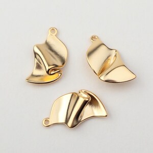 Puede incluir: Tres colgantes dorados de forma abstracta. Cada colgante tiene un lazo en la parte superior para sujetarlo a una joya. Los colgantes tienen un diseño plegado en forma de cinta con bordes curvos y un acabado brillante. Los colgantes están sobre un fondo blanco.