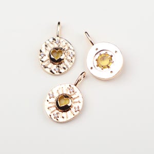 May include: Three gold-toned metal charms with circular designs. Two charms feature a central heart-shaped cutout surrounded by radiating lines and small clear stones. The third charm has a smooth border and a central yellow circular stone.