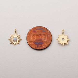 May include: Two gold-colored sunburst pendants with a central opal-like stone. The pendants are displayed next to a one-cent coin for size comparison. The pendants have a small loop at the top for hanging.