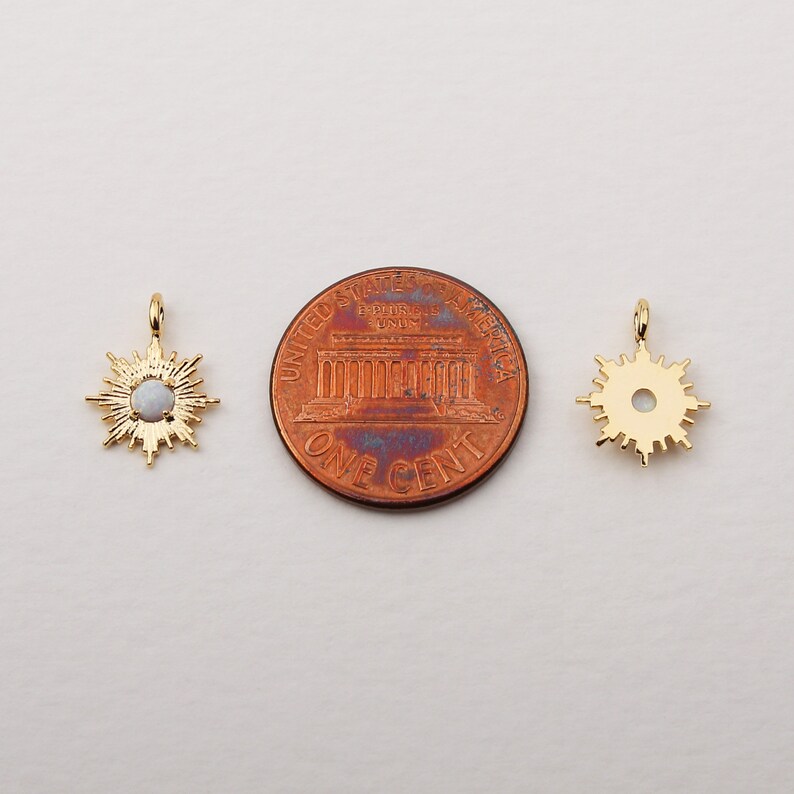 May include: Two gold-colored sunburst pendants with a central opal-like stone. The pendants are displayed next to a one-cent coin for size comparison. The pendants have a small loop at the top for hanging.