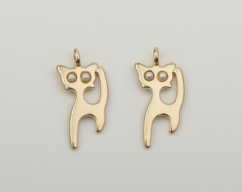 May include: Two gold-colored cat charms with white pearl eyes. The charms are in the shape of a cat standing on its hind legs with its tail curled around its body.