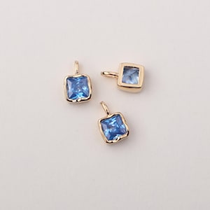 May include: Three gold-toned square charms with blue gemstone accents. The charms have a slightly textured edge.