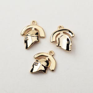 Puede incluir: Tres colgantes de metal dorado con forma de casco romano. Cada colgante tiene un lazo en la parte superior para sujetarlo a una cadena o pulsera. Un colgante tiene una pequeña gema transparente en el casco.