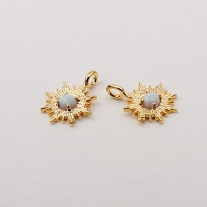 May include: Two gold-colored sunburst pendants with a central, round, opalescent stone. Each pendant has a small loop at the top for attaching to a chain or other jewelry component. The pendants are displayed against a plain, light background.