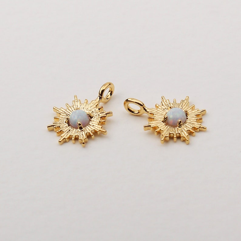 May include: Two gold-colored sunburst pendants with a central, round, opalescent stone. Each pendant has a small loop at the top for attaching to a chain or other jewelry component. The pendants are displayed against a plain, light background.