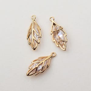 May include: Three gold-coloured leaf-shaped charms with clear crystal accents. Each charm has a loop at the top for attaching to jewellery. The leaves have detailed veining and a sparkling, faceted crystal centre.