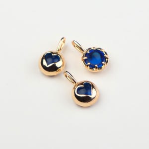 May include: Three small gold-toned metal charms with blue accents. Two charms feature a heart-shaped cutout revealing a deep blue center, while the third charm is a round setting containing a faceted blue stone with a pronged metal border.