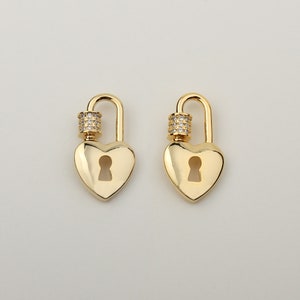 May include: Two gold-toned heart-shaped padlocks featuring a central keyhole cutout. Each padlock has a curved shackle adorned with a small cylindrical component encrusted with clear, sparkling cubic zirconia stones, creating a polished finish.