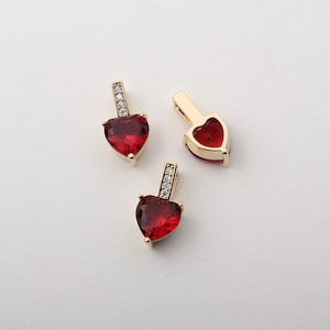 May include: Three gold-colored heart-shaped pendants with a vibrant red gemstone center. Each pendant has a small rectangular bail adorned with clear, sparkling stones. The pendants are displayed on a white surface.