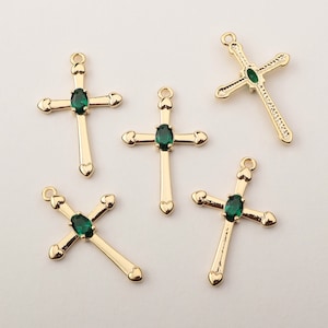 May include: Five gold-colored cross pendants with a green oval gemstone in the center. The crosses have heart-shaped details at the ends of the arms and a loop at the top for hanging. The background is a light, neutral color.