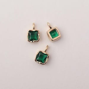 May include: Three gold-colored pendants with square, emerald-green gemstones. Each pendant has a small loop at the top for attaching to a chain or other jewelry component. The gemstones are set in a gold-colored bezel.