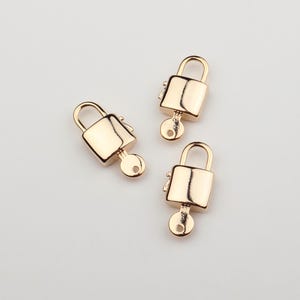 May include: Three small, gold-toned metal padlock charms, each featuring a smooth, rectangular body with a rounded shackle at the top and a small, integrated key shape protruding from the bottom center, finished with a polished, reflective surface.