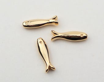 May include: Three small, gold-colored fish-shaped charms. Each charm has a smooth, reflective surface and a detailed tail fin. The charms are arranged on a plain, light-colored background, creating a simple and elegant presentation.