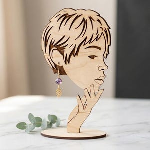 May include: Wooden earring display stand shaped like a woman's profile. The stand holds a pair of earrings with a purple gemstone and a gold leaf charm. The stand is made of light-colored wood.