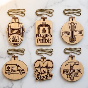 Może przedstawiać: Sześć drewnianych ozdób z różnymi grawerowanymi wzorami i tekstem. Wzory obejmują maskę spawalniczą z napisem "LET'S WELD", kran z napisem "FLUSHED WITH PRIDE" i termometr z napisem "KEEPING IT COOL". Każda ozdoba ma metalowy łańcuszek.