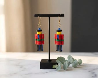 Puede incluir: Un par de pendientes hechos a mano con forma de soldados de juguete. Los pendientes presentan detalles en rojo, azul, verde, amarillo y negro. Cuelgan de ganchos dorados. Se exhiben en un soporte negro.