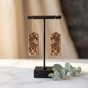 May include: Wooden earrings with detailed floral patterns. The earrings, with a light brown base and darker brown detailing, are rectangular and suspended from gold-coloured hooks. They are displayed on a black stand.