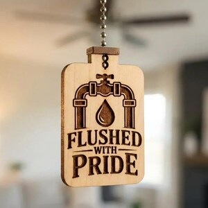 May include: A wooden ceiling fan pull featuring a laser-etched design. The design includes a water droplet, pipes, and the words "Flushed With Pride". The pull is attached to a silver chain.