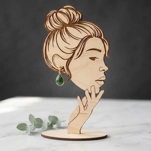 Peut inclure: Présentoir à boucles d'oreilles en bois en forme de profil de femme avec un chignon. Le présentoir contient une boucle d'oreille dorée avec une pierre verte en forme de goutte. Le présentoir est en bois clair et possède une base ronde.