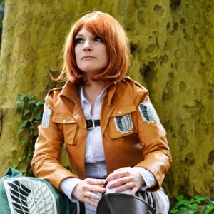 May include: A tan leather jacket with a black belt and white shirt. The jacket has two pockets and patches on the sleeves. The person is wearing a red wig and is sitting in front of a tree.