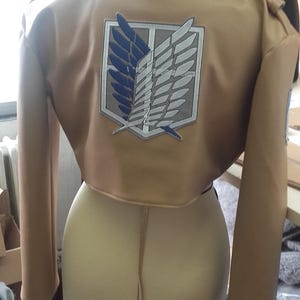 May include: Tan cropped jacket with long sleeves and a high collar. The back features a large emblem with blue and white wings within a white and gray border. The jacket is displayed on a mannequin.