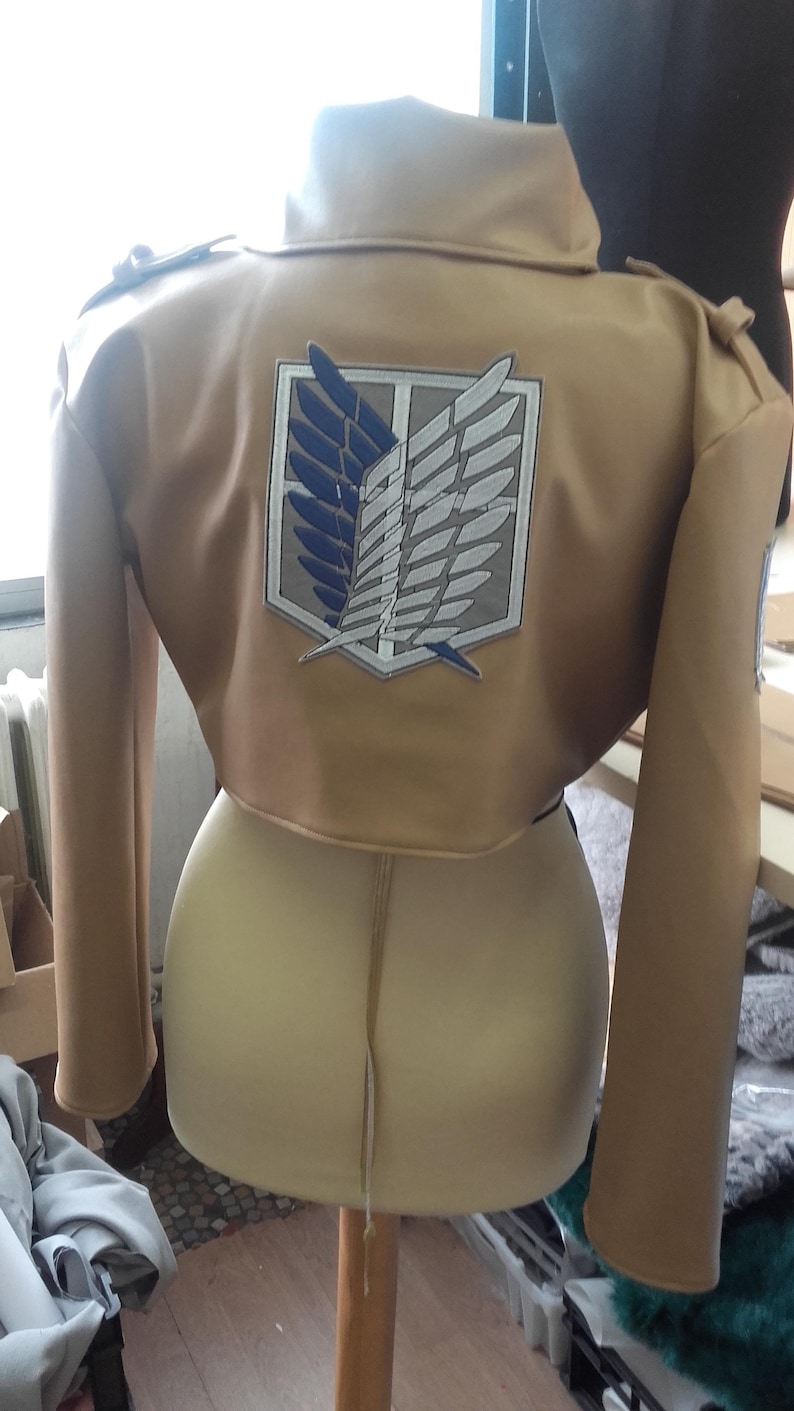 May include: Tan cropped jacket with long sleeves and a high collar. The back features a large emblem with blue and white wings within a white and gray border. The jacket is displayed on a mannequin.