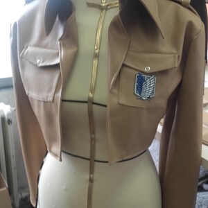 May include: A tan cropped jacket with a notched collar, two front pockets, and a patch with a winged emblem. The jacket is displayed on a mannequin. A gold-colored zipper runs down the front.