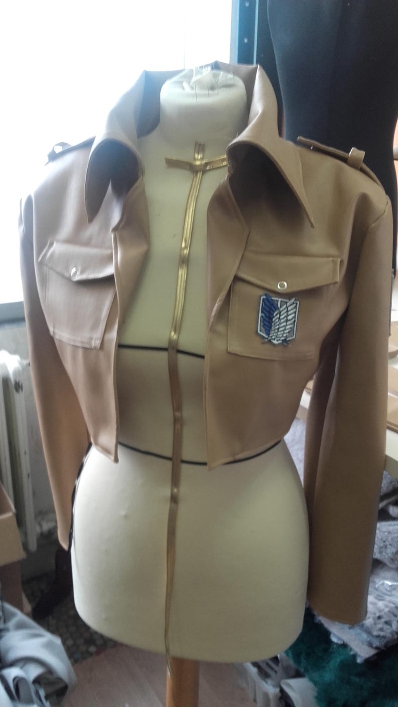May include: A tan cropped jacket with a notched collar, two front pockets, and a patch with a winged emblem. The jacket is displayed on a mannequin. A gold-colored zipper runs down the front.