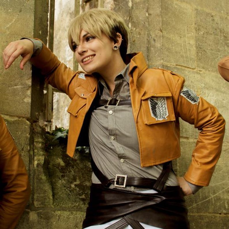May include: A tan leather jacket with multiple pockets and a cropped design. The jacket features a gray shirt underneath, a black belt, and a brown skirt. A patch with a wing design is visible on the jacket.