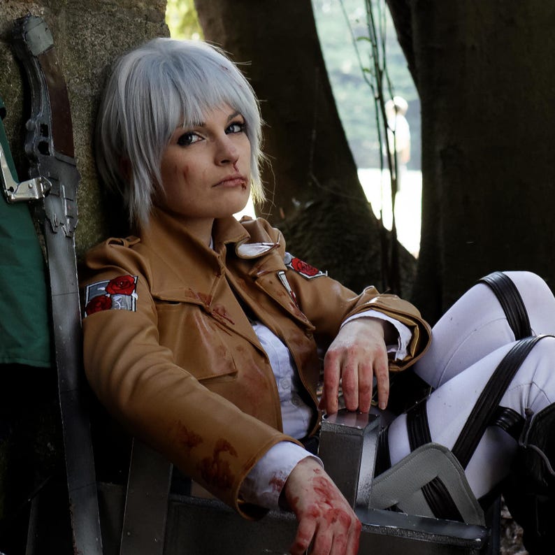 May include: A person in a brown jacket with red and white patches, white pants with black straps, and a short gray wig. The person is sitting in a chair with a sword leaning against a stone wall. The person has fake blood on their face and hands.