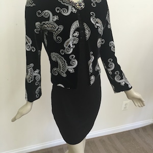 May include: A black cardigan with a silver paisley pattern and a jeweled brooch. The cardigan has long sleeves and a v-neck.