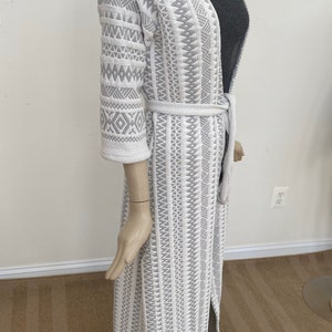 May include: A long white cardigan with a gray geometric pattern. The cardigan has a tie belt and a long sleeve.