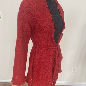 Puede incluir: Una chaqueta de punto de encaje roja con cuello festoneado y cinturón de lazo. La chaqueta se muestra en un maniquí.