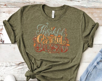 May include: Olive green t-shirt with the words "Thankful, Grateful, Blessed" in a colorful, decorative font. The shirt is paired with light blue jeans and white sneakers, suggesting a casual, everyday outfit. The design includes fall-themed leaf accents.