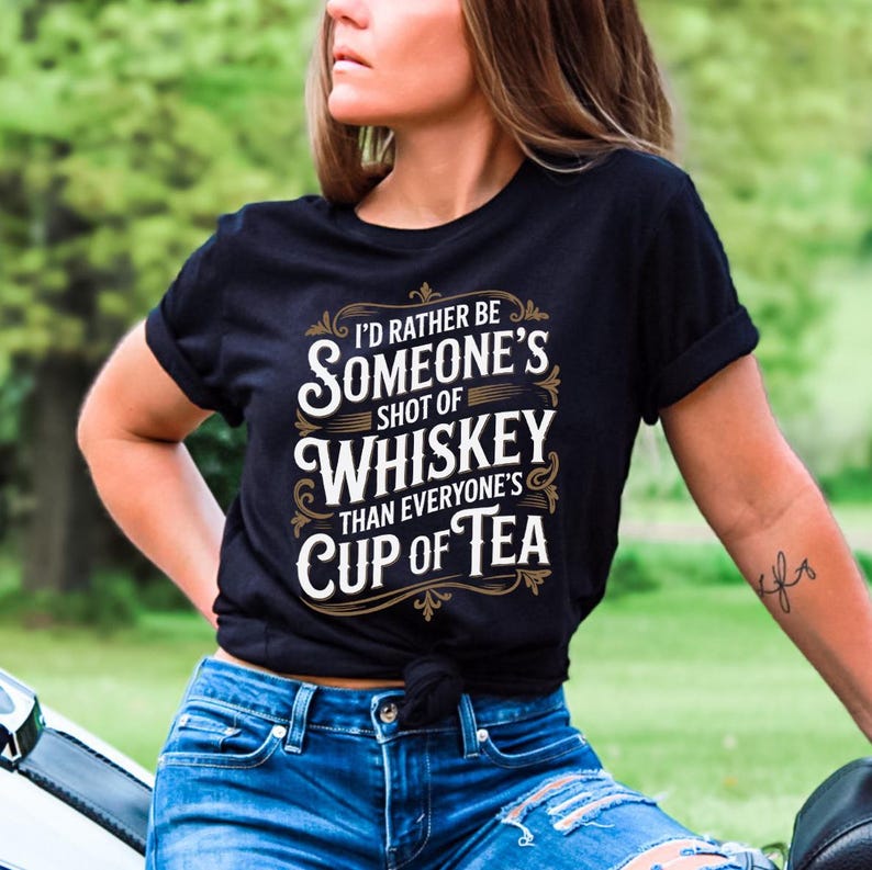 May include: Black t-shirt with the text "I'd Rather Be Someone's Shot of Whiskey Than Everyone's Cup of Tea" in white and gold lettering. The shirt is worn by a person in blue jeans.