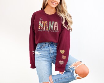May include: Burgundy sweatshirt with the text "The Nana" in colorful patterned letters and the text "wearing her heart on her sleeve" in white. The sleeves have three small hearts with names on them.