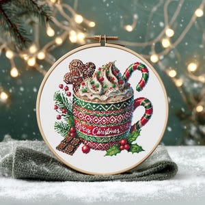 May include: A cross-stitch design of a festive holiday mug filled with whipped cream, a candy cane, and a gingerbread cookie. The mug is decorated with a red and green pattern and the words "Merry Christmas". The design is set in a wooden embroidery hoop.