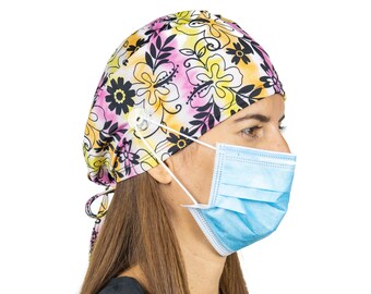 May include: A woman wearing a pink, yellow, and black floral print surgical scrub hat with a white button and a blue surgical mask.