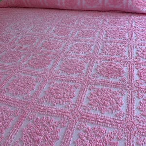 May include: A light pink bedspread featuring a repeating square grid pattern. Each square contains a raised, textured floral motif in the center. The bedspread has a matching bolster pillow at the top, also displaying the same pink floral design.