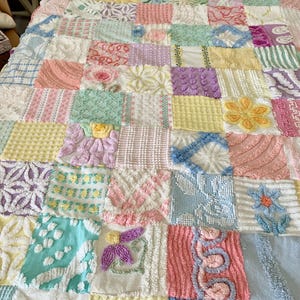 May include: A patchwork quilt featuring a grid of square fabric blocks in pastel shades of pink, blue, yellow, green, and white. Each square displays unique raised textures, including floral motifs, geometric patterns, and tufted chenille designs.
