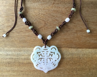 Op de afbeelding: Een ketting met een witte jade hanger met een gegraveerd tijgerontwerp. De ketting is gemaakt van bruin koord en groene en witte kralen.