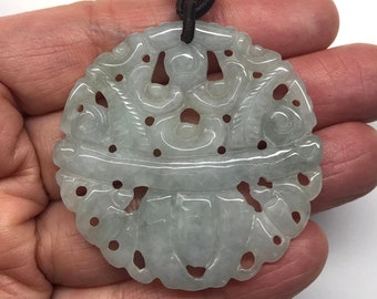 Op de afbeelding: Een jade ketting met een ronde, ingewikkeld gegraveerde jade hanger. De hanger heeft een delicaat bloemenontwerp met opengewerkt details. De ketting is gemaakt van bruin koord en groene jade kralen.