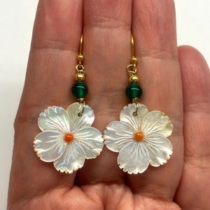 May include: A pair of gold earrings with a white mother of pearl flower charm and a green glass bead. The flower has a small orange center.