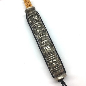 May include: A silver prayer wheel with intricate carvings and a black cord. The prayer wheel is decorated with a repeating pattern of symbols and figures.