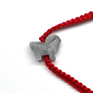 May include: A bracelet featuring a bright red woven cord and a single, carved light gray stone charm shaped like a ram. The cord is constructed with a repeating knotted pattern, and the stone charm is positioned centrally on the red string.